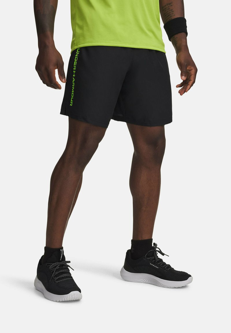 Pantaloni sportivi neri con logo "Under Armour" verde, abbinati a sneaker neri e una maglietta verde lime. I pantaloni hanno una texture liscia.