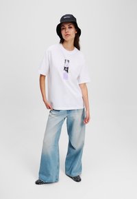 White short-sleeve T-shirt with a graphic print and lavender accent, paired with wide-leg light blue jeans and black bucket hat.