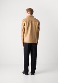 Άνδρας με ταμπά δερμάτινο μπουφάν, μαύρο φαρδύ παντελόνι και μαύρα παπούτσια, που στέκεται μπροστά σε έναν λευκό τοίχο με τα χέρια του στα πλάγια.