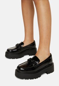Sorte lakkert skinn loafers med tykke plattformer og dekorativt tilbehør. Glatt tekstur med høyglanset finish og rund tådesign.