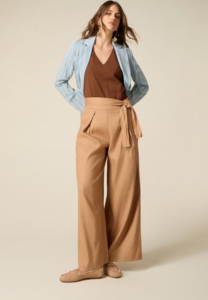 Kvinde står iført beige bukser med vide ben og bindebånd i taljen, brun V-hals top, lyseblå stribet blazer og beige mokkasiner.