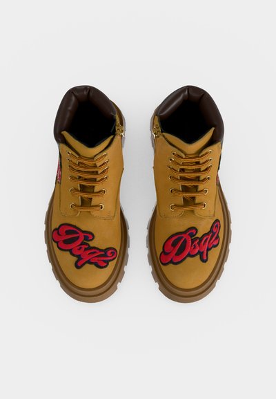 Paire de bottes en cuir fauve avec col matelassé marron, lacets jaunes, semelles épaisses et écussons rouges "Dsq2" sur le dessus avant de chaque chaussure.
