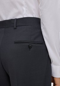 Biała koszula włożona w ciemnoszare spodnie garniturowe z pojedynczą zapinaną kieszonką na plecach, widoczną pętelką na pasek i prawą ręką wzdłuż ciała.