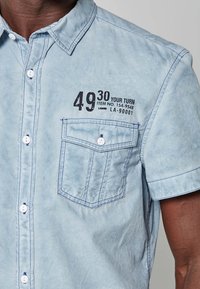 Vaaleansininen lyhythihainen paita, jossa on etupuolella napitettu tasku, valkoiset napit, tummat saumat ja musta painettu teksti "49 30 YOUR TURN" rinnan kohdalla.