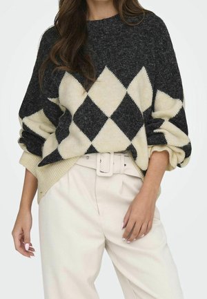 Femme portant un pull noir et crème à motif losanges avec des manches bouffantes, rentré dans un pantalon crème taille haute avec une ceinture assortie.