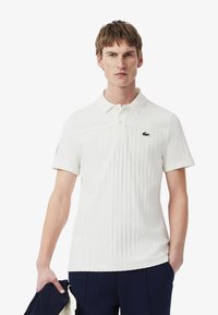 Vit pikétröja med vertikal ribbad textur, tre knappar och en grön logotyp på vänster bröst. Bärs med marinblå byxor.