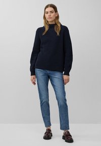 Marineblauwe ribgebreide trui met hoge hals, gecombineerd met lichtblauwe denim jeans en donkerburgundy instappers, op een neutrale achtergrond.