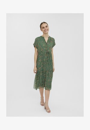 Femme souriante marchant vers l'avant dans une robe midi verte fleurie avec des boutons, des manches courtes et des talons blocs beige à bride cheville sur fond blanc.