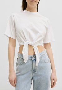 Witte cropped t-shirt met korte mouwen, voorzien van een geknoopt voorkantdetail. Gecombineerd met lichtblauwe high-waisted jeans.