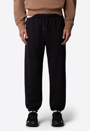 Personne portant un pantalon de survêtement noir The North Face avec des poignets élastiques et des baskets noires, debout devant un fond clair uni.