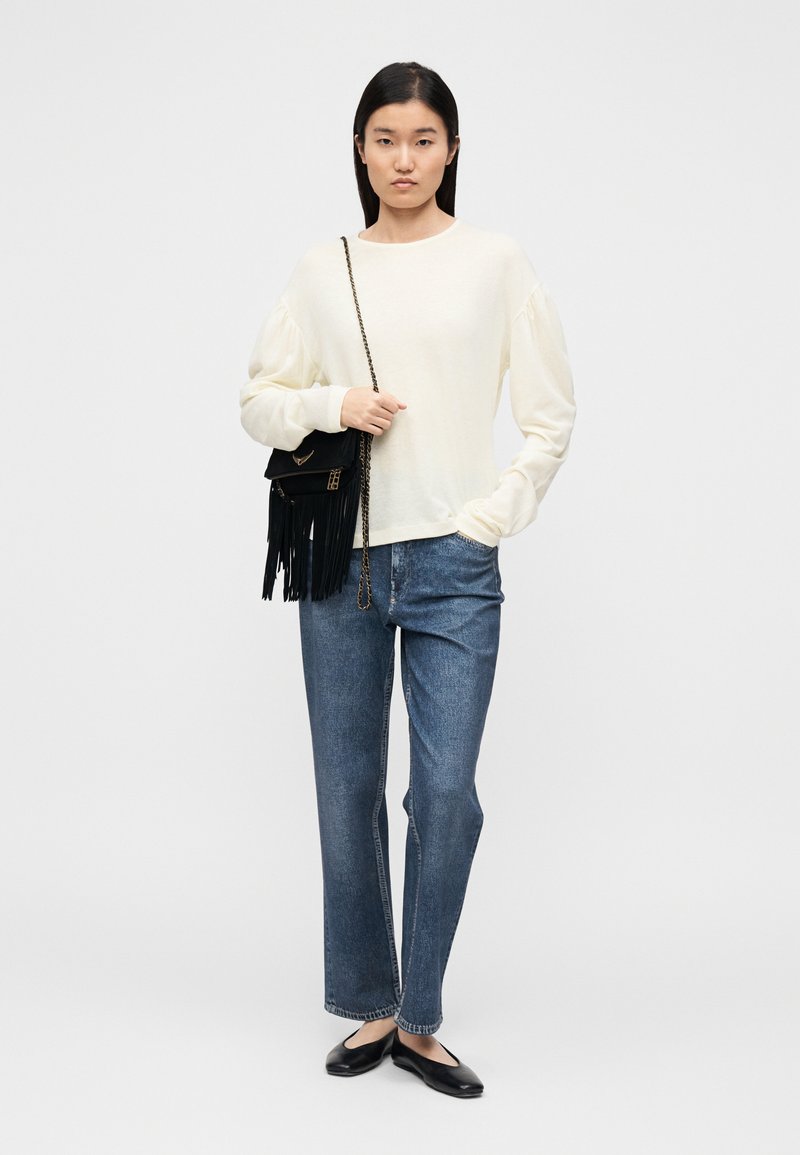 Pull couleur crème à manches bouffantes, associé à un jean droit bleu. Sac bandoulière noir avec détails à franges et chaussures plates noires.