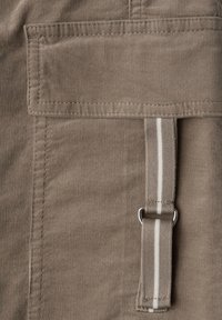 Close-up van een bruine stoffen zak met klep en verticale gestreepte band bevestigd met een metalen D-ring op een gestructureerd materiaal.