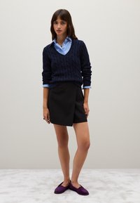 Jersey de punto trenzado azul marino sobre una camisa de botón de color azul claro, combinado con una falda envolvente negra y unos zapatos de escaso en color púrpura.