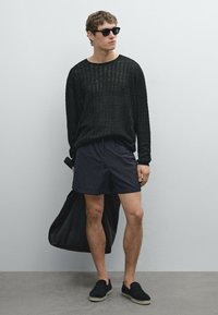 Černý pletený svetr, tmavé šortky, espadrilky, sluneční brýle, drží černou tašku. Neutrální pozadí; zaměření na oblečení a doplňky.