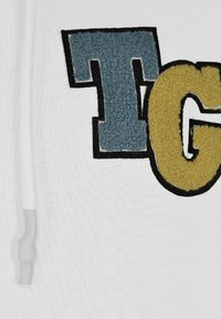 Vit sweatshirt med broderade bokstäver "T" i blått frottétyg och "G" i gult, omgiven av svart, med dragsko i halsringningen.