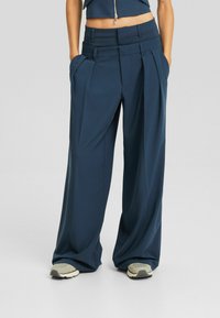 Marinblå vida byxor i slätt tyg med framveck, djupa fickor och hög midja, kombinerat med ljusgrå sneakers.
