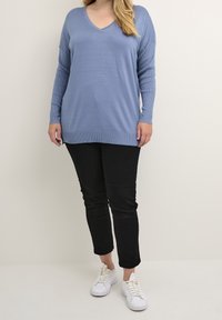 Jasnoniebieski sweter z dekoltem w serek, wykonany z dzianiny, z długimi rękawami, ściągaczami przy nadgarstkach i u dołu, zestawiony z czarnymi spodniami slim-fit i białymi trampkami.