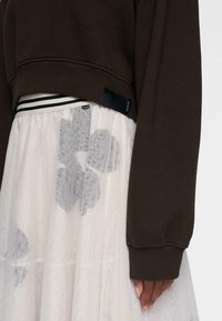 Hånden hviler ved siden iført en mørkebrun langærmet bluse og en hvid nederdel med sorte abstrakte blomstermønstre under et gennemsigtigt stof.