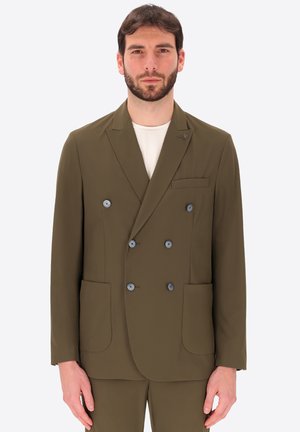 Uomo che indossa blazer doppiopetto verde oliva con bottoni grigi e tasche applicate, in piedi su sfondo bianco semplice.