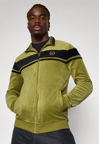 Green velvet track jacket with black stripes, golden logo, zip closure, side pockets, and ribbed cuffs and hem. Textured, soft surface.