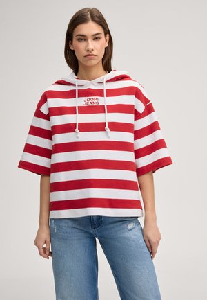 Jonge vrouw die een rood-witte kortärmelig hoodie met horizontale strepen en de tekst "JOOP! JEANS" draagt, gecombineerd met lichtblauwe jeans met beschadigde details.