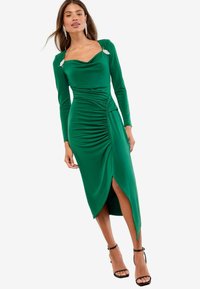 Rochie verde, cu mâneci lungi, cu decolteu în formă de cowl, cu un corset plisat, fente lateral și accente decorative din metal pe umeri. Material neted, cu o cădere fluidă.