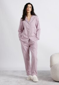 Γυναίκα όρθια με τα χέρια στις τσέπες, φοράει ροζ και λευκή πιτζάμα με καρό σχέδιο και λευκά παντόφλες σε γκρίζο χαλί δίπλα σε μια λευκή καρέκλα.