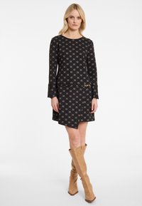 Blonde Frau, die ein schwarzes, gemustertes Kleid mit langen Ärmeln und goldenen Verschlussdetails trägt, steht vor einem weißen Hintergrund. Sie trägt auch beige kniehohe Stiefel.