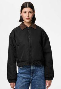 Kobieta ubrana w czarną pikowaną kurtkę z brązowym kołnierzem z korduroju i niebieskie dżinsy, stojąca na tle jednolitego białego tła.