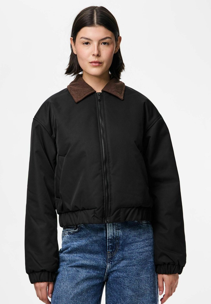 Kobieta ubrana w czarną pikowaną kurtkę z brązowym kołnierzem z korduroju i niebieskie dżinsy, stojąca na tle jednolitego białego tła.