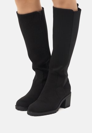Wysokie czarne zamszowe kozaki na blokowym obcasie noszone na nogach, z bocznymi suwakami i elastycznymi panelami.