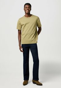 T-shirt verde oliva a maniche corte con scollo rotondo, abbinato a jeans blu scuro e stivali marroni. Tessuto morbido, vestibilità casual.