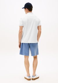 Man in een marineblauwe pet, wit poloshirt met korte mouwen, blauwe shorts en witte sneakers die met zijn rug naar de camera staat.