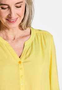 Blusa gialla leggera con scollo a V, cinque bottoni frontali, dettagli arricciati sulle spalle e una texture morbida.