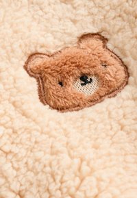 Plush blanket with a soft, cream texture featuring an embroidered bear face in tan, with black and beige accents.
