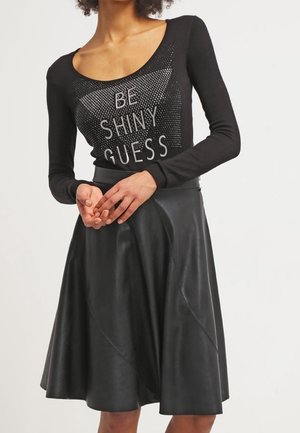 Kobieta ubrana w czarną bluzkę z długim rękawem z napisem "BE SHINY GUESS" oraz czarną spódnicę o długości do kolan w kształcie litery A z subtelnymi przeszyciami.