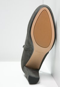 Grå högklackad sko i mocka med texturerad yta, triangulär tå och en brun gummisula med en diskret logotyp.