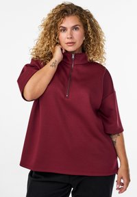 Frau mit lockigem blonden Haar, die ein bordeauxfarbenes Half-Zip-Oberteil und schwarze Hosen trägt, und ihren Hals berührt, während ein Tattoo auf ihrem Unterarm sichtbar ist.