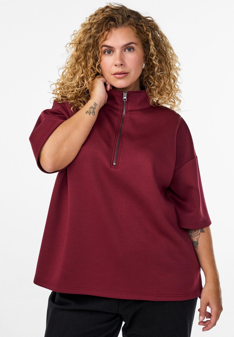 Frau mit lockigem blonden Haar, die ein bordeauxfarbenes Half-Zip-Oberteil und schwarze Hosen trägt, und ihren Hals berührt, während ein Tattoo auf ihrem Unterarm sichtbar ist.