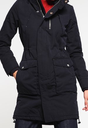 Žena v dlouhém černém zimním kabátu s předními kapsami s klopami a detaily na zipu, stojící s jednou rukou v kapse před jednobarevným pozadím.