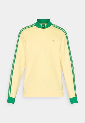Dzeltena džemperis ar zaļiem akcentiem, ar pusi rāvējslēdzēju, augstu apkakli un zaļām trīssvītru detaļām gar piedurknēm. Mazs logo uz krūšu.