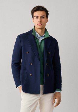 Hombre con blazer azul marino de doble botonadura sobre jersey verde con cremallera y camisa a rayas, combinado con pantalones beige claro, de pie sobre fondo liso.