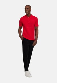 Polo shirt rossa con colletto, maniche corte e tre bottoni, abbinata a pantaloni neri e sneakers bianche su uno sfondo neutro.