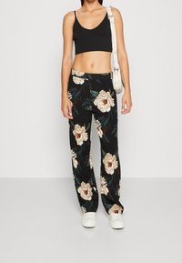 Kobieta ubrana w czarny crop top, czarne kwiatowe szerokie spodnie, białe trampki i białą torebkę na ramię, stojąca na jednolitym tle.