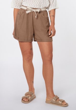 Person trägt braune Shorts mit hoher Taille und weißem gebundenem Gürtel, ein weißes Stricktop und beige gewebte Slip-On-Sandalen.
