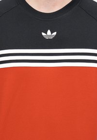 T-shirt de manga curta vermelho e preto com três riscas horizontais brancas e um pequeno logótipo branco. Material em algodão macio, design com gola redonda.