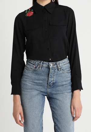 Femme portant une chemise noire boutonnée avec une broderie florale rouge sur l'épaule et un jean bleu clair taille haute.