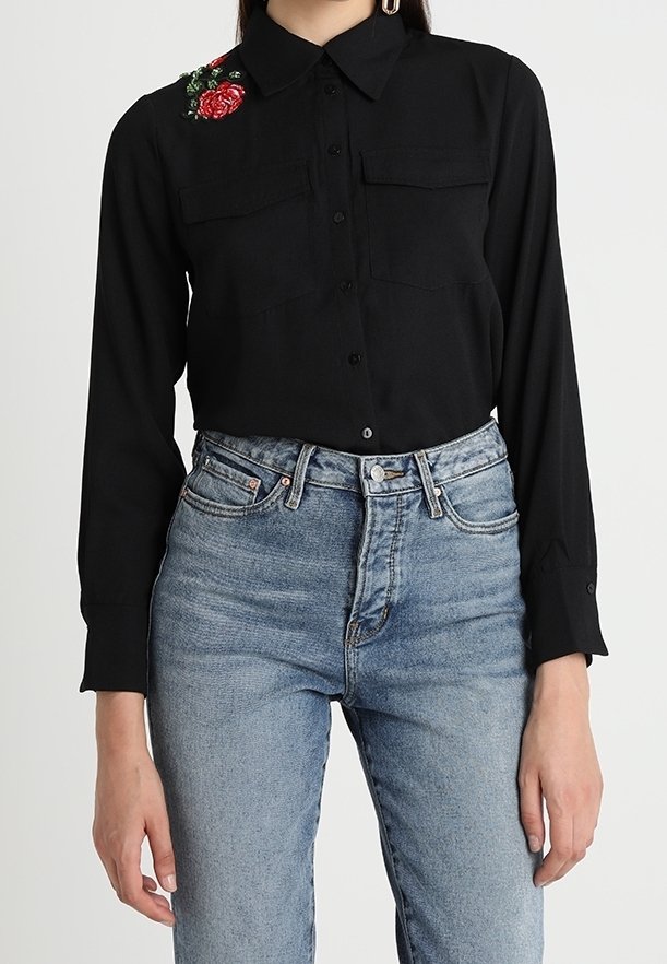 Femme portant une chemise noire boutonnée avec une broderie florale rouge sur l'épaule et un jean bleu clair taille haute.