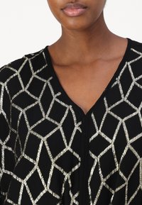 Svart tyg topp med ett geometriskt mönster i metallic silver, v-ringning och texturerad yta.