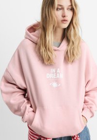 Jeune femme avec de longs cheveux blonds portant un sweat à capuche rose pastel avec le texte "DANS UN RÊVE" et les mains dans la poche avant.